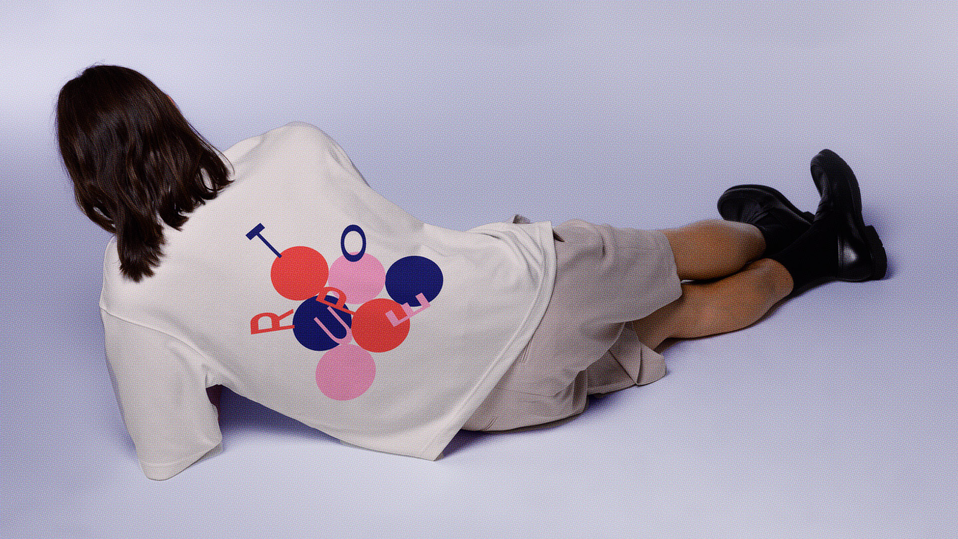 A figure lays on her side, showing the back of her t-shirt with Troupe letters balancing and falling down a stack of circles in red, blue, and pink.